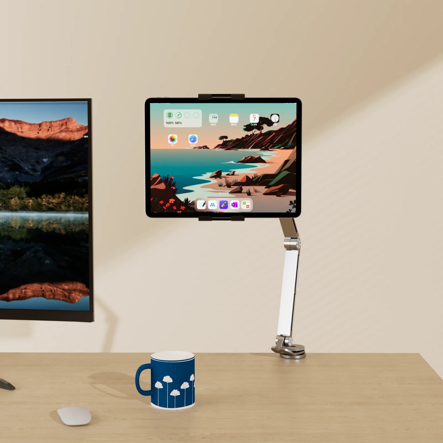 An Artpowers Adjustable Tablet Holder mounts a tablet displaying a colorful landscape next to a computer monitor on a wooden desk. A blue mug with white clouds and a computer mouse are also present.