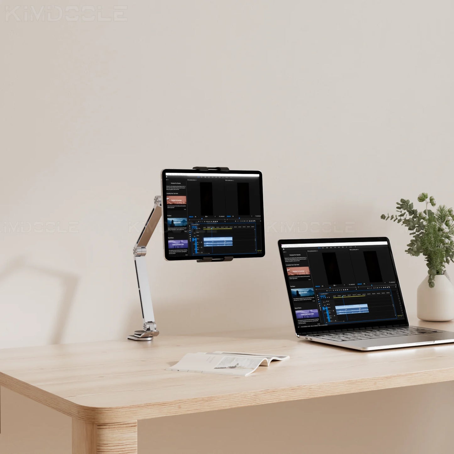 A minimalist workspace showcases a tablet on the Artpowers Adjustable Tablet Holder - Versatile Stand, and a laptop on a wooden desk, both displaying video editing software. A small green plant adds natures touch, while an open notebook lies ready for notes.