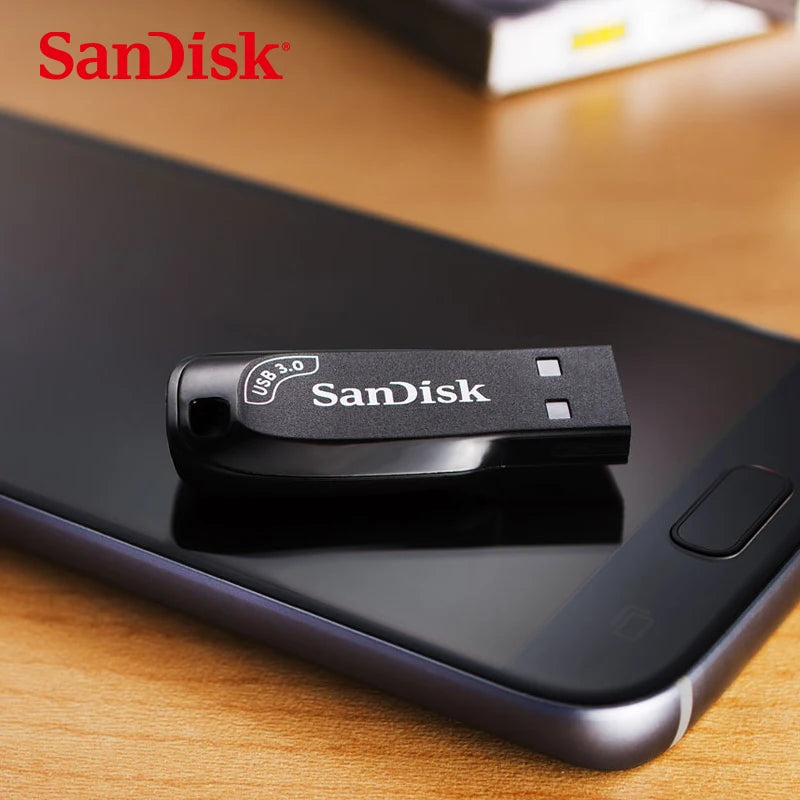 A black SanDisk USB 3.0 Flash Drive - High-Speed Compact Storage rests on a smartphone atop a wooden surface. The device, featuring a visible button, showcases the prominent SanDisk logo on both the drive and in the images top left corner.