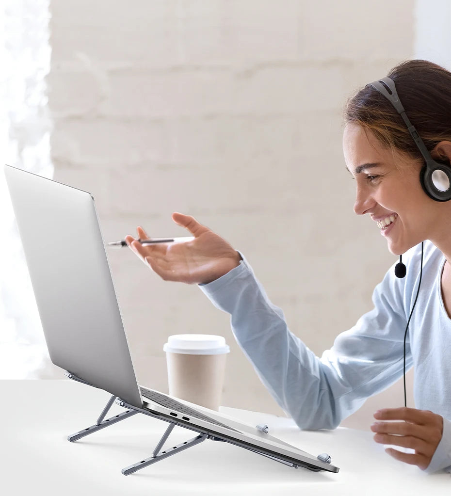 Smiling during a video call, a person with a headset uses an UGREEN Laptop Stand - Adjustable Foldable Aluminum for their laptop. They gesture with a pen, sitting at a table with take-out coffee against a soft, bright, and relaxed background.