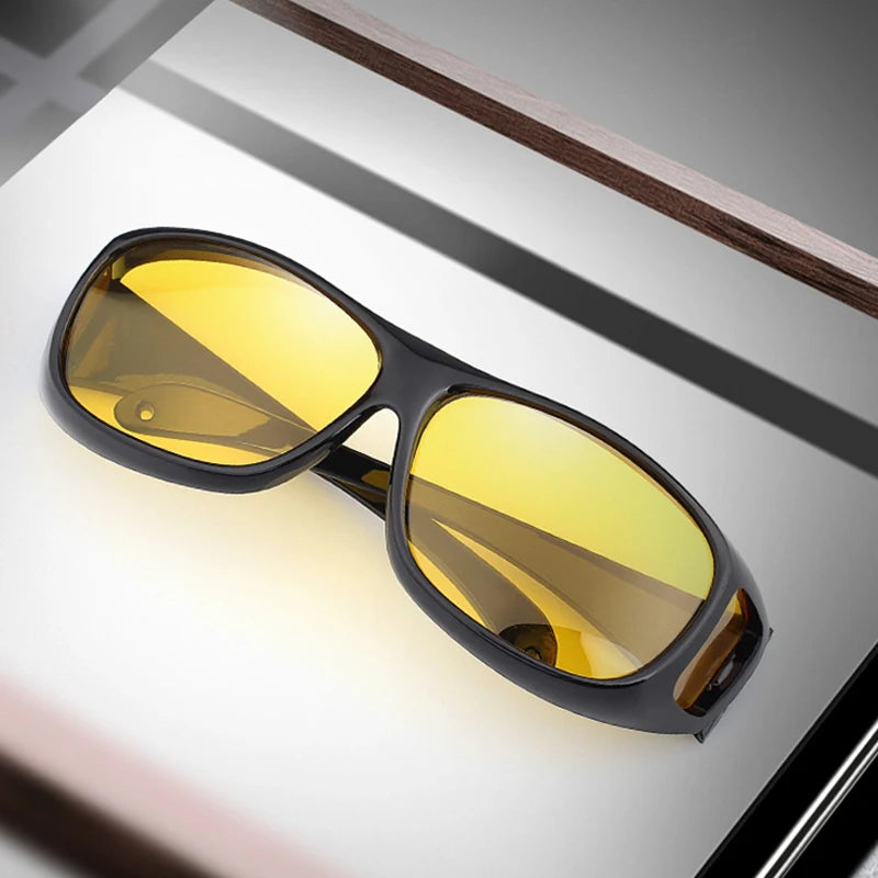 NoEnName Night Vision Glasses with yellow-tinted lenses and a black frame rest on a reflective surface, angled to display both lenses. Perfect for driving, the background features subtle brown and gray tones.