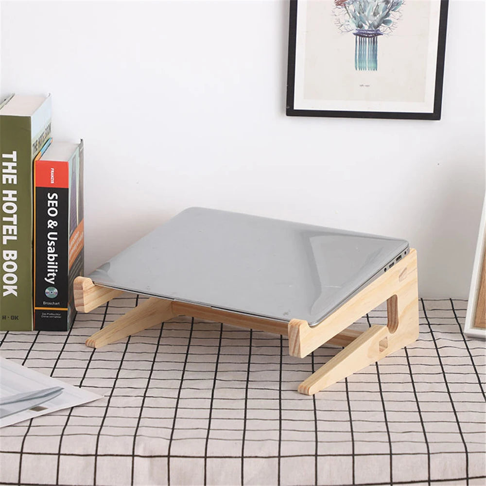 The Universal Wooden Laptop Holder - Detachable Stand cradles a closed laptop on a checkered tablecloth with books upright to the left. A framed vase picture adds charm to the background. Enjoy free shipping on this elegant stand.