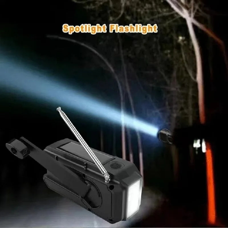 The Sayin Portable Solar Radio with Power Bank & Flashlight shines a bright beam into a dark forest, illuminating tree branches. Featuring a black, rectangular design, foldable handle, and built-in power bank, this emergency tool ensures youre ready for any adventure.