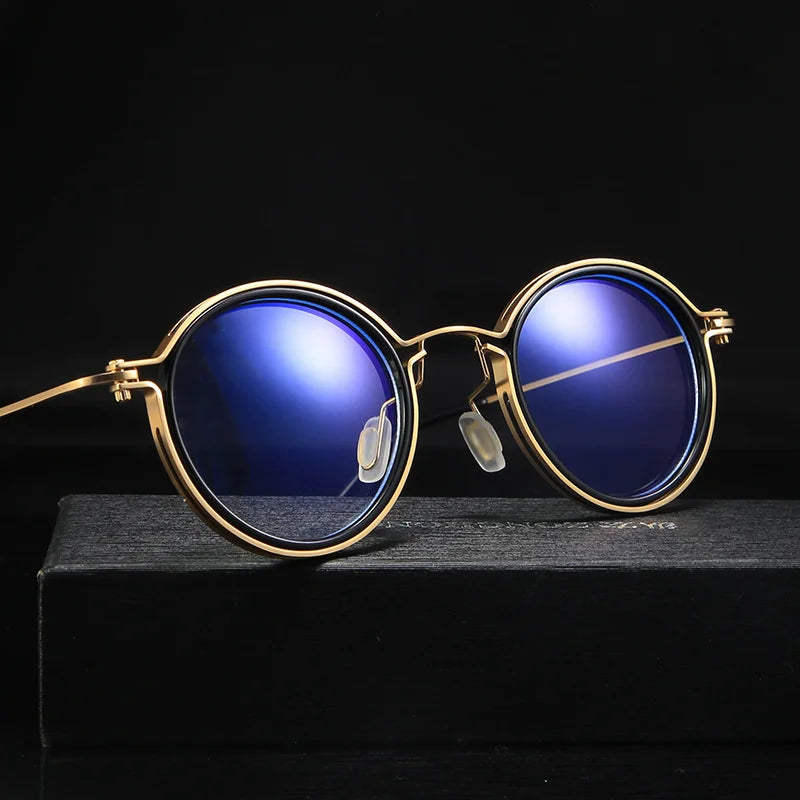KLASSNUM Luxury Round Frame Reading Glasses feature anti-blue light reflective blue lenses in a gold metal frame. They sit on a black surface against a dark background, with thin temples adding sleekness. Perfect for style and protection on sunny days, their round frames offer timeless appeal.