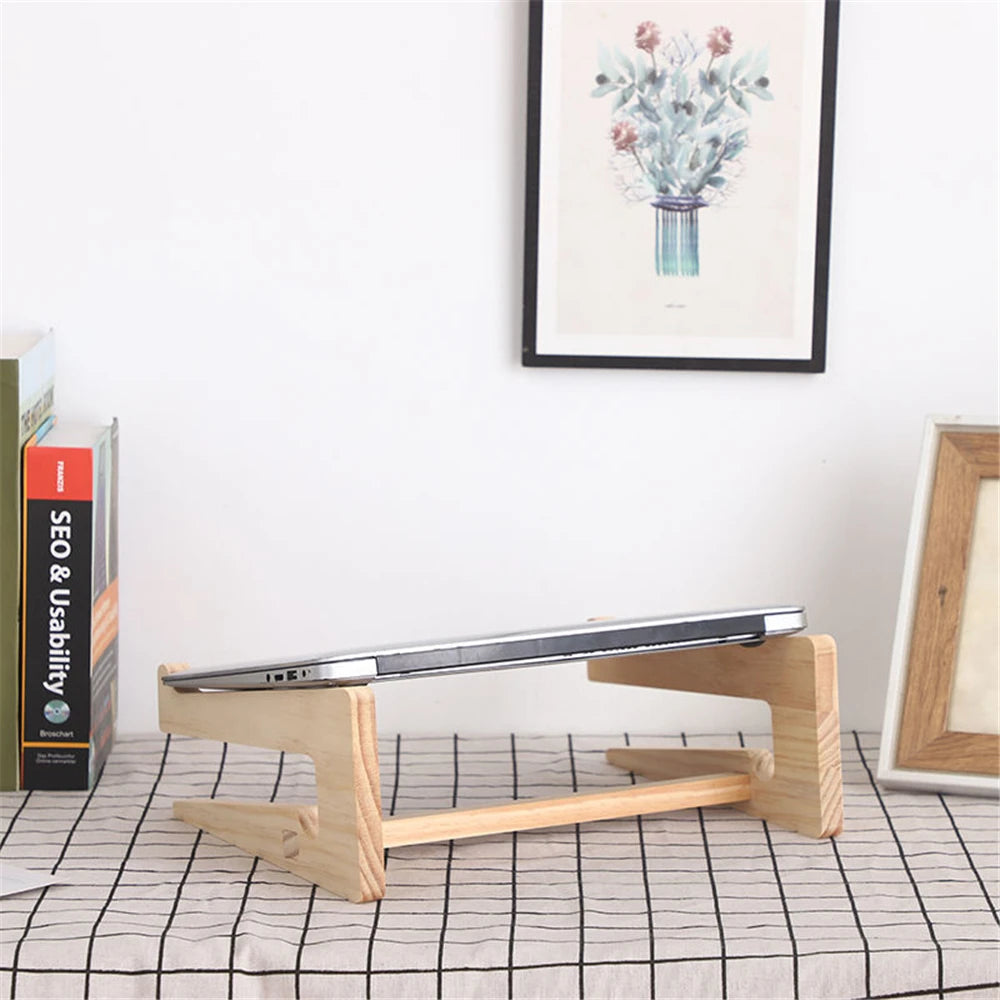 The Universal Wooden Laptop Holder - Detachable Stand tilts your closed laptop elegantly on your desk, complemented by books on the left and framed floral artwork on a white wall, with another frame to the right. Enjoy free shipping while elevating the style and function of your workspace.