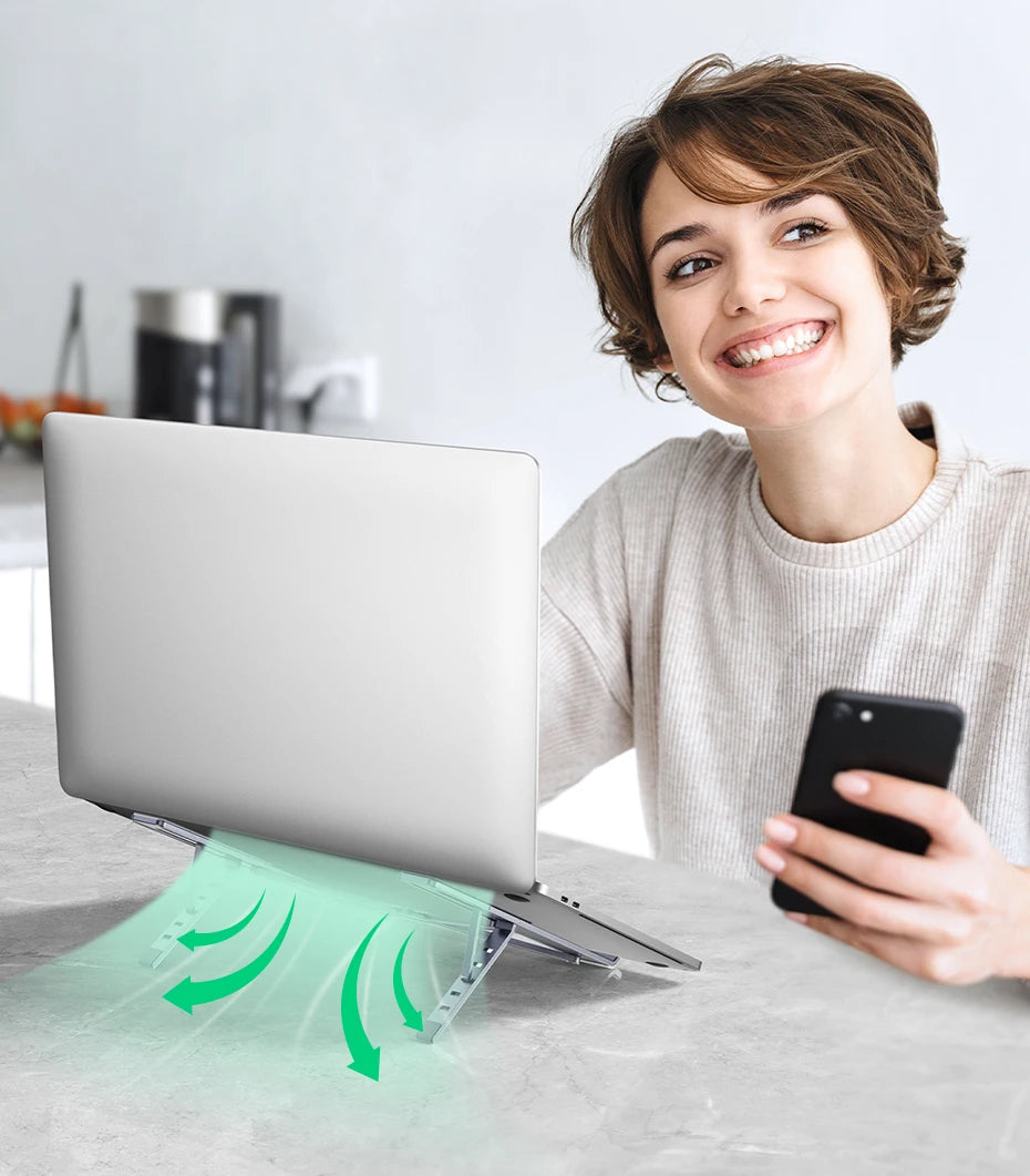 A person smiles at a table using a smartphone, with their laptop on the UGREEN Laptop Stand - Adjustable Foldable Aluminum. Green arrows under the laptop show airflow. A blender and kitchen items are in the background.
