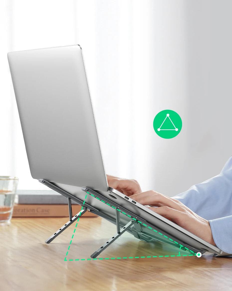 A person types on a laptop raised by the UGREEN Laptop Stand - Adjustable Foldable Aluminum, highlighted with a green triangle overlay. The ergonomic setup rests on a wooden table, with a clear glass and book nearby.