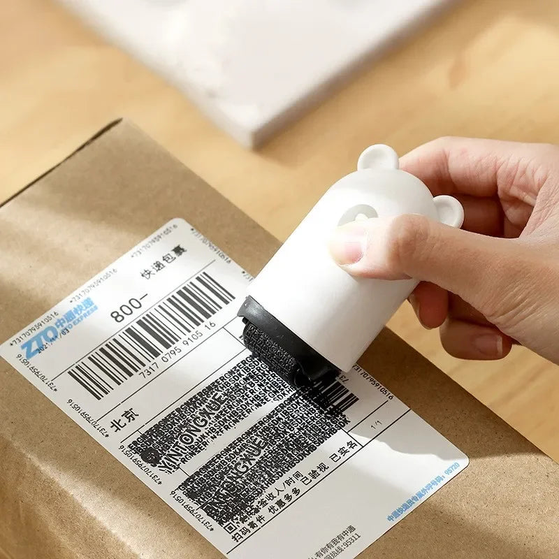 A hand uses the NoEnName Roller Stamp - Quick Drying Privacy Seal, featuring a cute white bear design, to quickly cover personal info on a shipping label attached to a brown cardboard box.