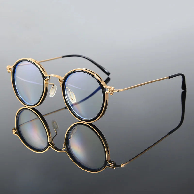KLASSNUM Mens Round Glasses feature stylish, round gold frames with retro charm and blue-tinted anti-blue light lenses. Thin temples with black tips and a clear bridge add modern flair, resting on a reflective surface that shows the underside of these fashionable glasses.