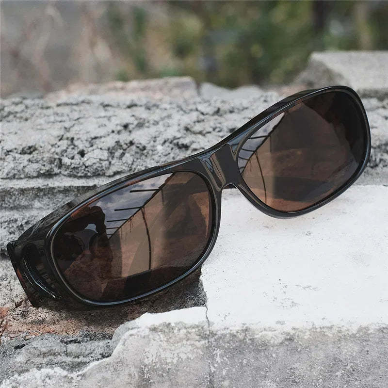 MOKDUFF Polarized Sunglasses, designed for comfortable wear over glasses, in black with wrap-around UV400 protection and dark lenses, are set on a rough rocky surface against a blurred natural background.