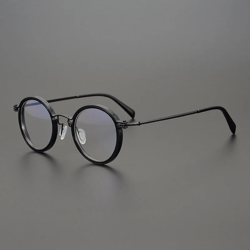 KLASSNUM Mens Round Glasses with retro black frames and anti-blue light lenses sit on a flat, dark gray surface. They have flexible nose pads and sleek temples. The subtle lens reflection adds to their simple, classic appeal, making them ideal mens eyewear.