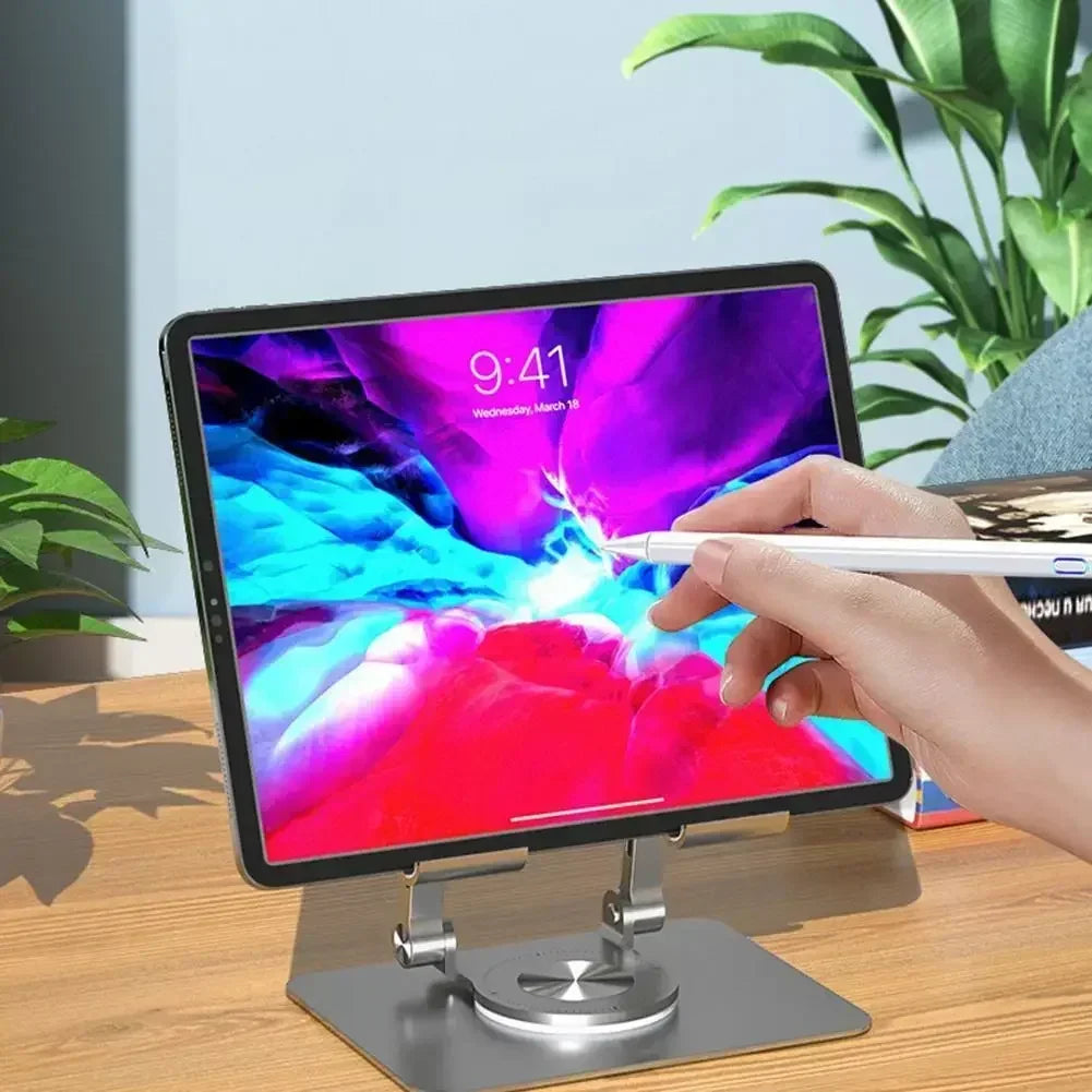 A tablet with a vibrant abstract display is on the Adjustable 360° Rotatable Tablet Stand for 4.7-12 Devices, placed atop a wooden desk. A hand uses a stylus on the screen, framed by surrounding plants and books that add greenery and color to the background.