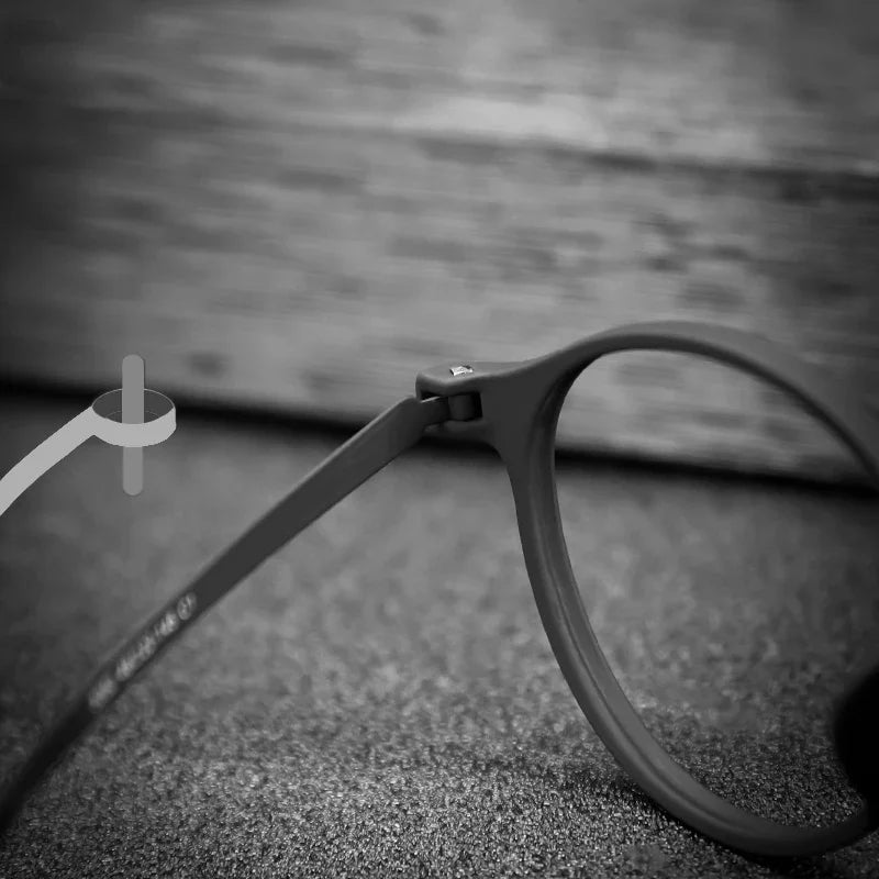 The black and white image showcases the OUTMIX Pure Titanium Anti Blue Light Reading Glasses resting on a textured surface, emphasizing the sleek titanium hinge and frame against a blurred background, which highlights its craftsmanship and elegance.