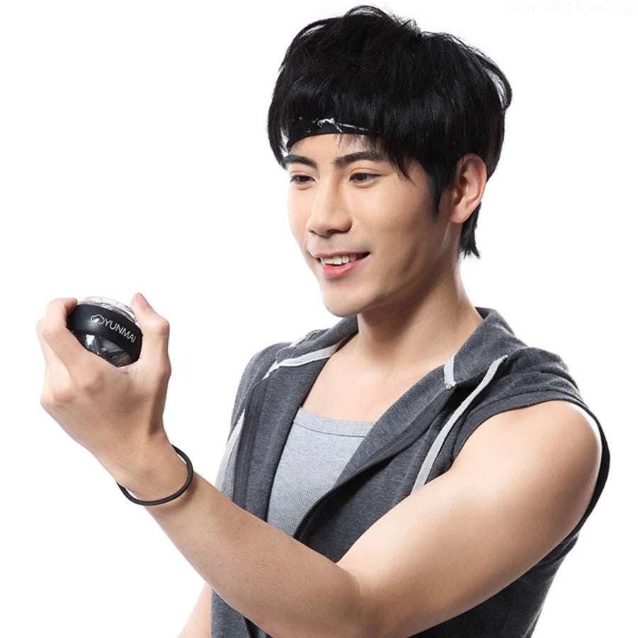 A person with short black hair and a headband smiles while holding a small black Xiaomi Forearm Exerciser LED Gyroball Trainer. They wear a sleeveless gray hoodie over a light gray shirt against a white background.
