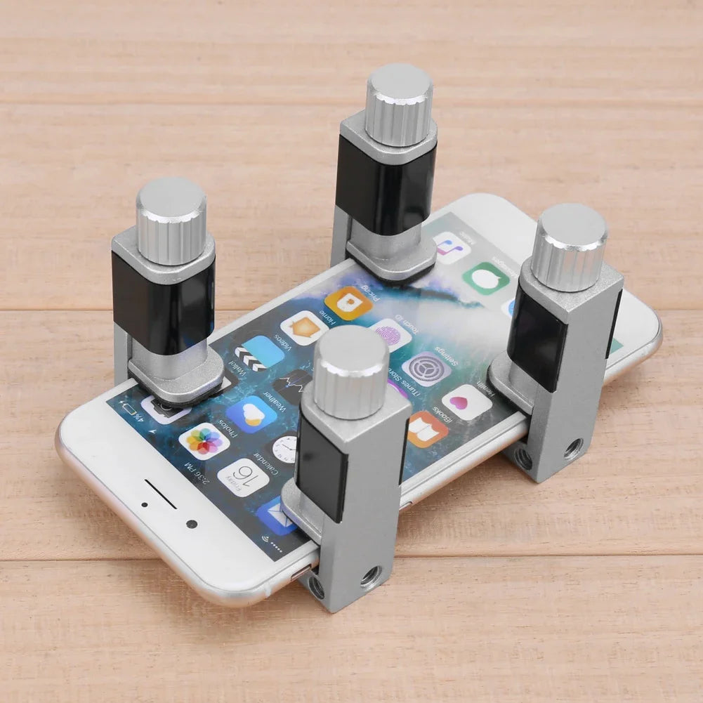 A smartphone lies face-up on a wooden surface with the MOJOYCE Adjustable Alloy Clamp for Secure Mobile Repairs placed on top, featuring four metal posts with adjustable clamps at the screens corners for safe repairs or optimal screen protection.