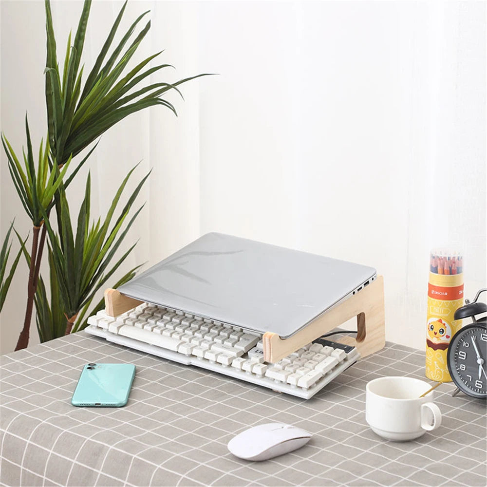 A Universal Wooden Laptop Holder raises a laptop above a keyboard on a table with a gray checkered cloth. Nearby are a smartphone, white mug, wireless mouse, plant, and colored pencils. Enjoy free shipping!.