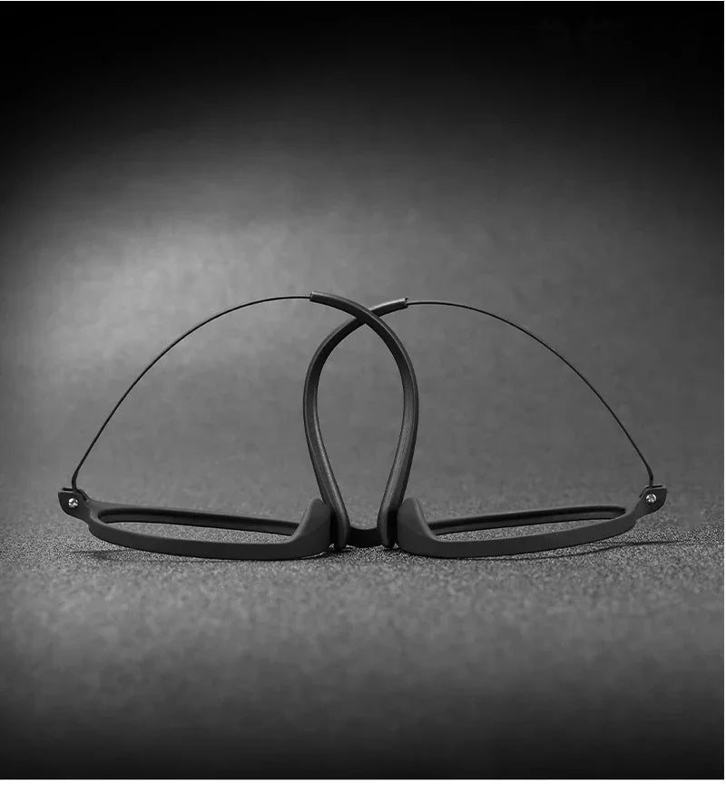The OUTMIX Pure Titanium Anti Blue Light Reading Glasses feature a sleek wire-like black titanium frame, folded and upright on a textured dark surface against a gradient gray background, creating an elegant arch for stylish sophistication.