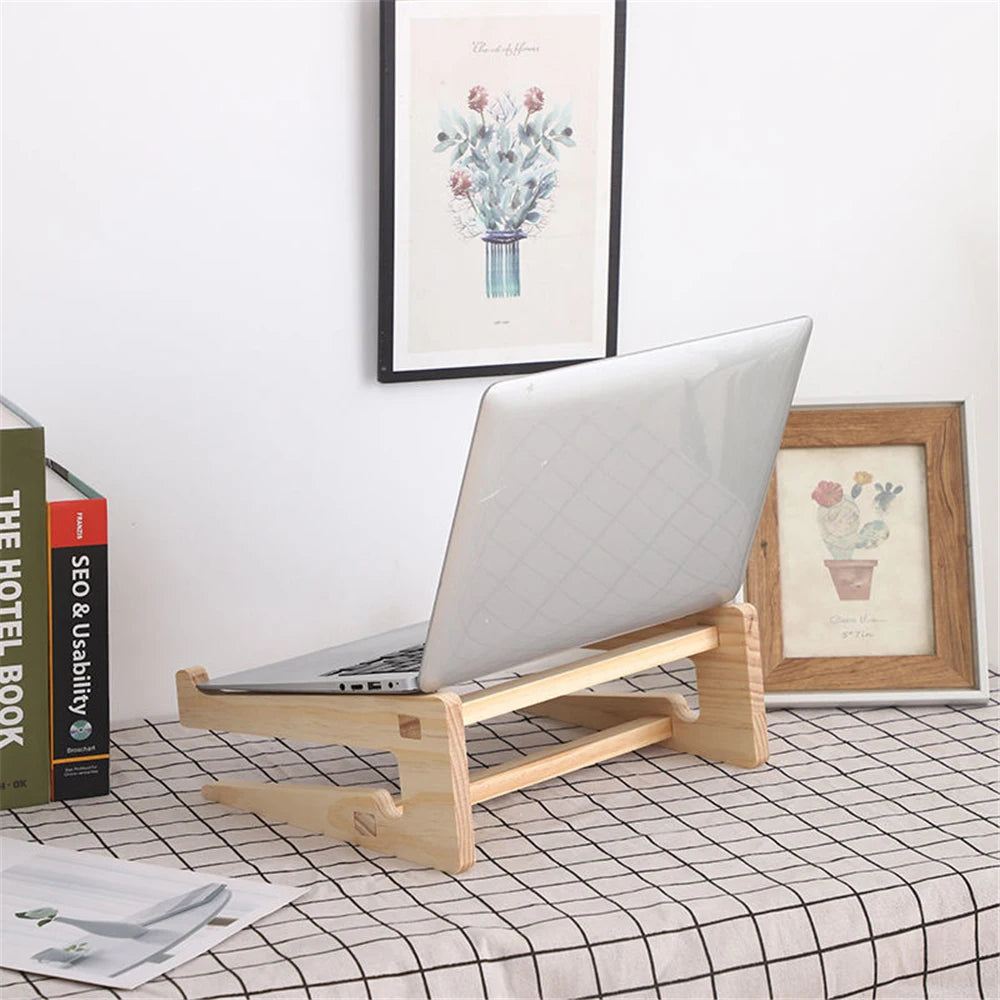 A laptop sits on a Universal Wooden Laptop Holder - Detachable Stand on a checkered tablecloth. The background features two framed artworks, one depicting a floral bouquet. Books, including an SEO guide, are stacked to the left. Free shipping is available for this arrangement.
