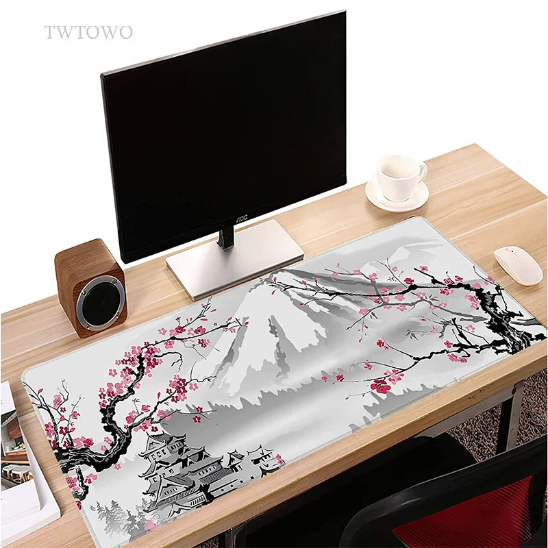 On the desk, a computer monitor, keyboard, and mouse accompany the TWTOWO Sakura XL Mouse Pad—adorned with serene pink cherry blossoms and featuring a non-slip base—with a white cup next to a small speaker.