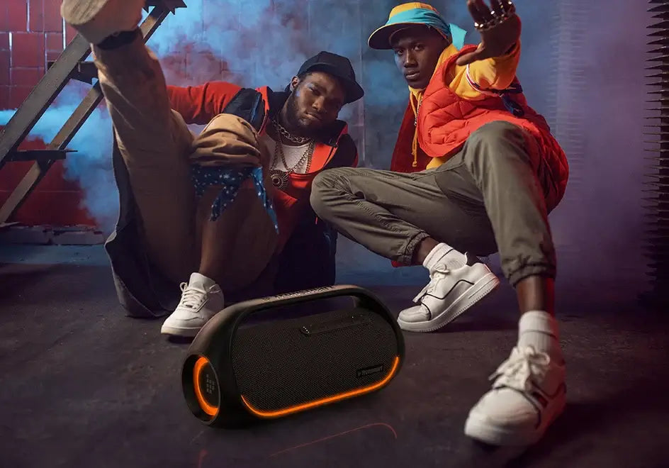 Two people pose stylishly with sneakers, crouching beside the Tronsmart Bang Speaker, a 60W Bluetooth device with illuminated edges. Dramatic lighting and smoke effects form a dynamic urban scene that promises to energize any atmosphere with its heavy bass.
