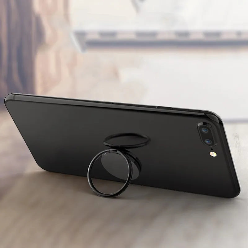 A smartphone on a table features a sleek Luxury Metal 360° Rotation Mobile Phone Ring Holder. Its dual camera and flash stand out, with the magnetic ring holder ensuring stable positioning against the blurred wall and ledge backdrop.