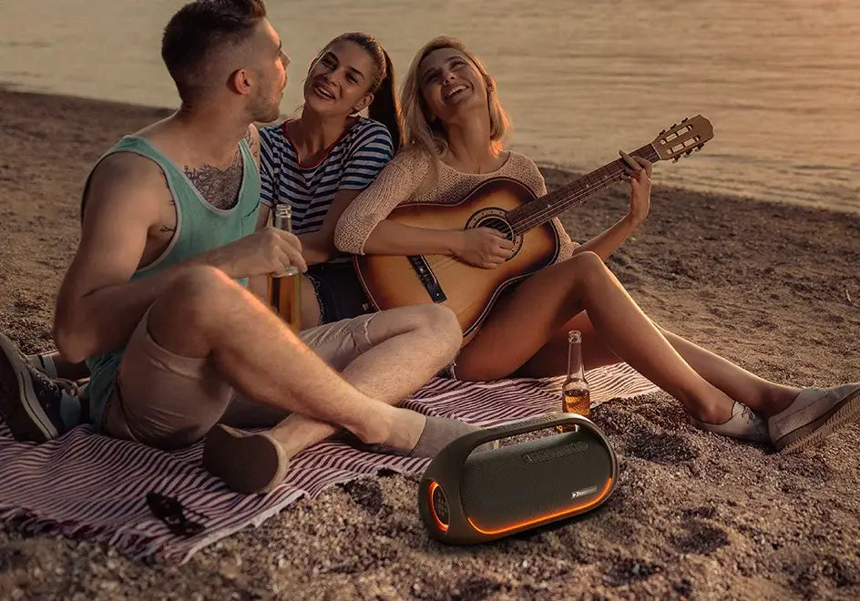 Three people relax on a beach during sunset. One plays the guitar while the others listen and smile, seated on a striped blanket with drinks nearby, as music from a Tronsmart Bang Speaker fills the air, its 60W Bluetooth heavy bass perfectly blending with app-controlled playlists.