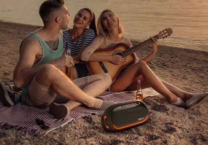 Three people relax on a beach during sunset. One plays the guitar while the others listen and smile, seated on a striped blanket with drinks nearby, as music from a Tronsmart Bang Speaker fills the air, its 60W Bluetooth heavy bass perfectly blending with app-controlled playlists.