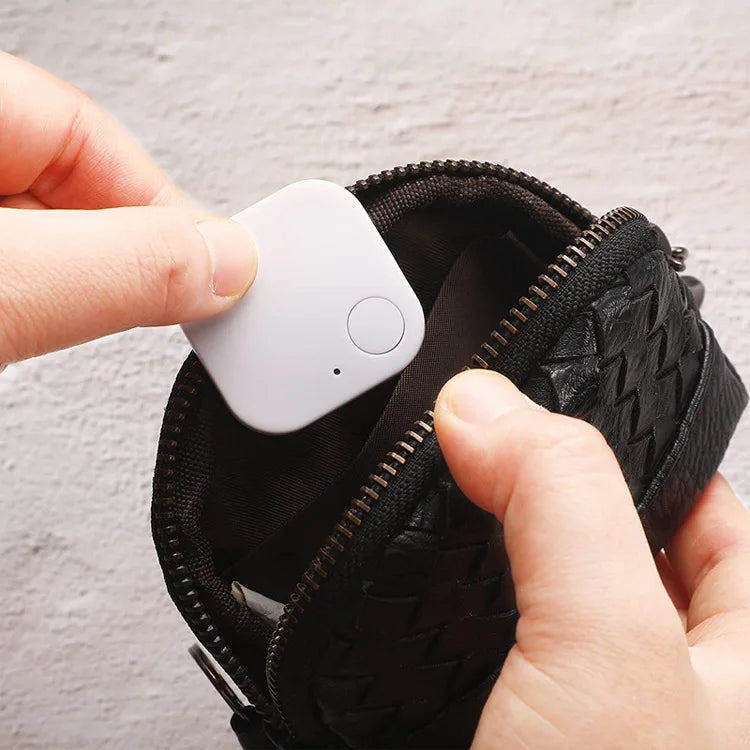 A person places a small, square white Smart Bluetooth 4.0 Anti-Lost Tracker for Valuables with a button into a sleek black zippered pouch featuring a woven texture, against a light background.