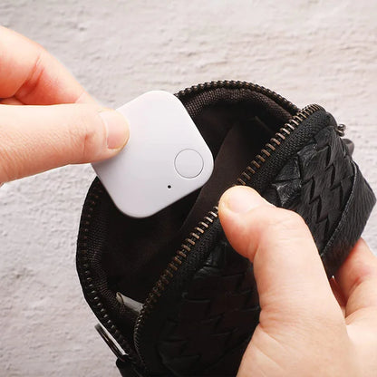 A person places a small, square white Smart Bluetooth 4.0 Anti-Lost Tracker for Valuables with a button into a sleek black zippered pouch featuring a woven texture, against a light background.