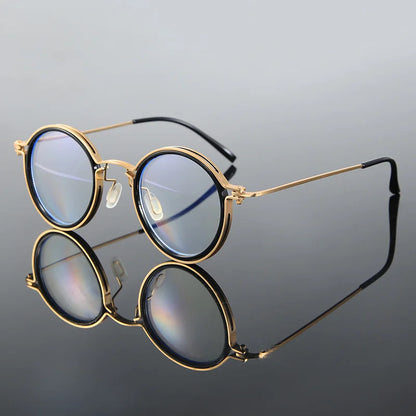 KLASSNUM Luxury Round Frame Reading Glasses, featuring gold metal frames and reflective lenses, are placed on a glossy surface that mirrors them. The glasses have thin earpieces with black tips, set against a grey gradient background.
