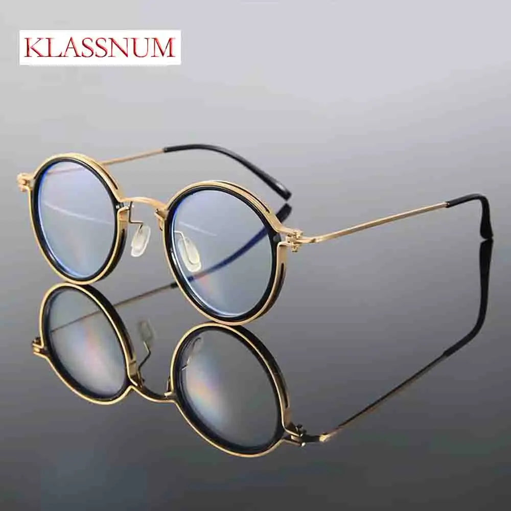 The KLASSNUM Mens Round Glasses feature gold frames and anti-blue light lenses, resting stylishly on a reflective black surface. The brand name is elegantly inscribed in red at the top left corner, highlighting their retro charm.
