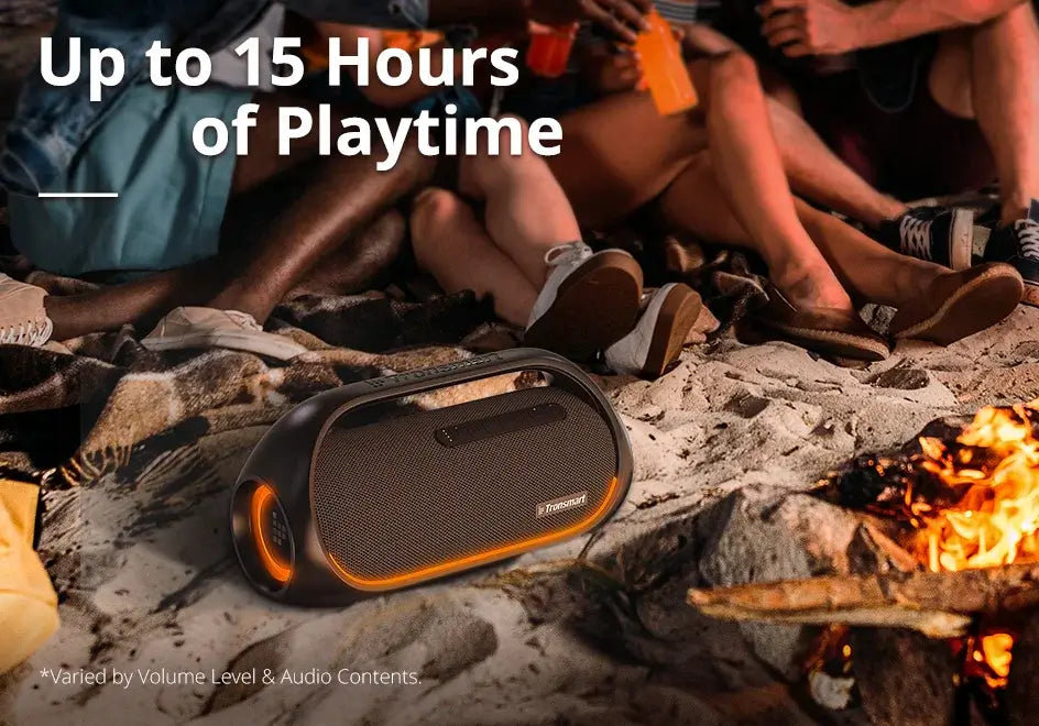 A Tronsmart Bang Speaker 60W Bluetooth with heavy bass and orange accents rests on the sand by a campfire. People enjoying drinks surround the fire as music plays, with up to 15 hours of playtime available.