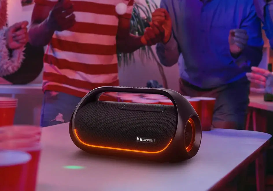 A Tronsmart Bang Speaker with 60W Bluetooth and heavy bass, accented by orange LED lights, energizes the room as people dance. Red plastic cups add to the lively party atmosphere. The colorful, dynamic vibe is easily controlled via an app for seamless enjoyment.