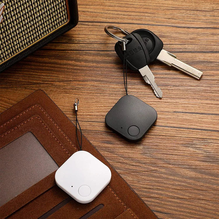 White and black Smart Bluetooth 4.0 Anti-Lost Trackers for Valuables rest on a wooden surface, accompanied by a set of keys with a black car key fob. A brown leather wallet peeks into the frame beside the black tracker.