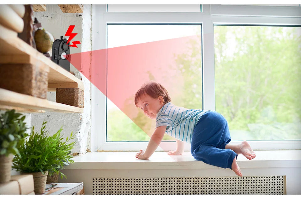 A baby climbs on a windowsill as the KERUI Mini Motion Detector Alarm on a shelf detects movement, activates its red alert light, and is ready to trigger its 120DB siren for added personal security.