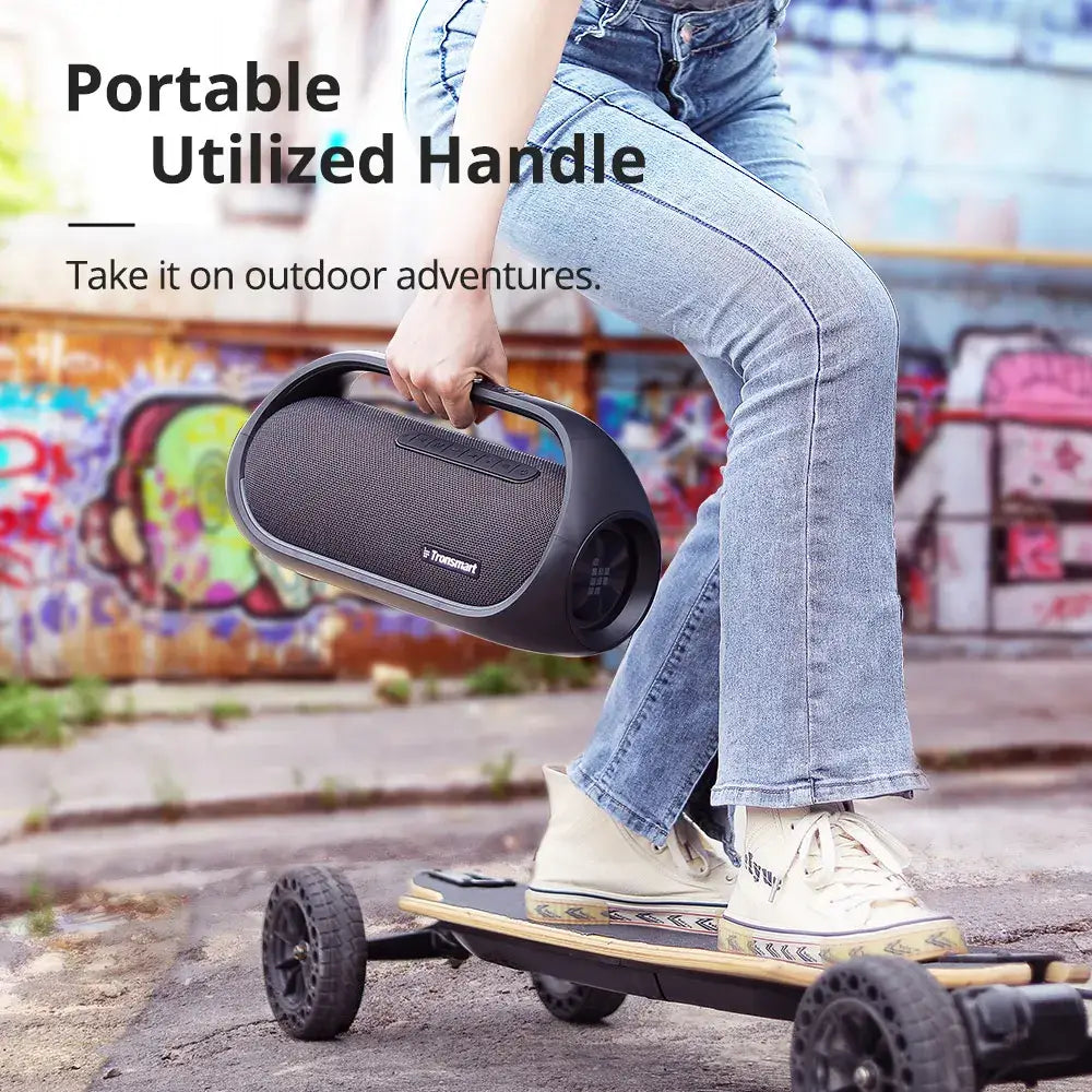 A person in jeans stands beside a skateboard with large wheels, holding the Tronsmart Bang Speaker 60W Bluetooth with a handle. The vibrant graffiti background complements the scene. Text reads: Portable with Heavy Bass & App Control—Perfect for Outdoor Adventures.