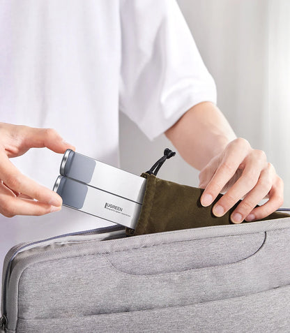 A person in a white shirt is placing the UGREEN Laptop Stand - Adjustable Foldable Aluminum, featuring an ergonomic design and gray accents, into a brown drawstring pouch inside a gray bag.