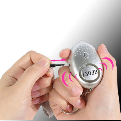 A hand grasps the 130dB Personal Safety Alarm Keychain, its pin pulled for self-defense. Emitting pink sound wave graphics to illustrate its intense alertness, it hangs from a keychain strap, offering peace of mind and personal safety.